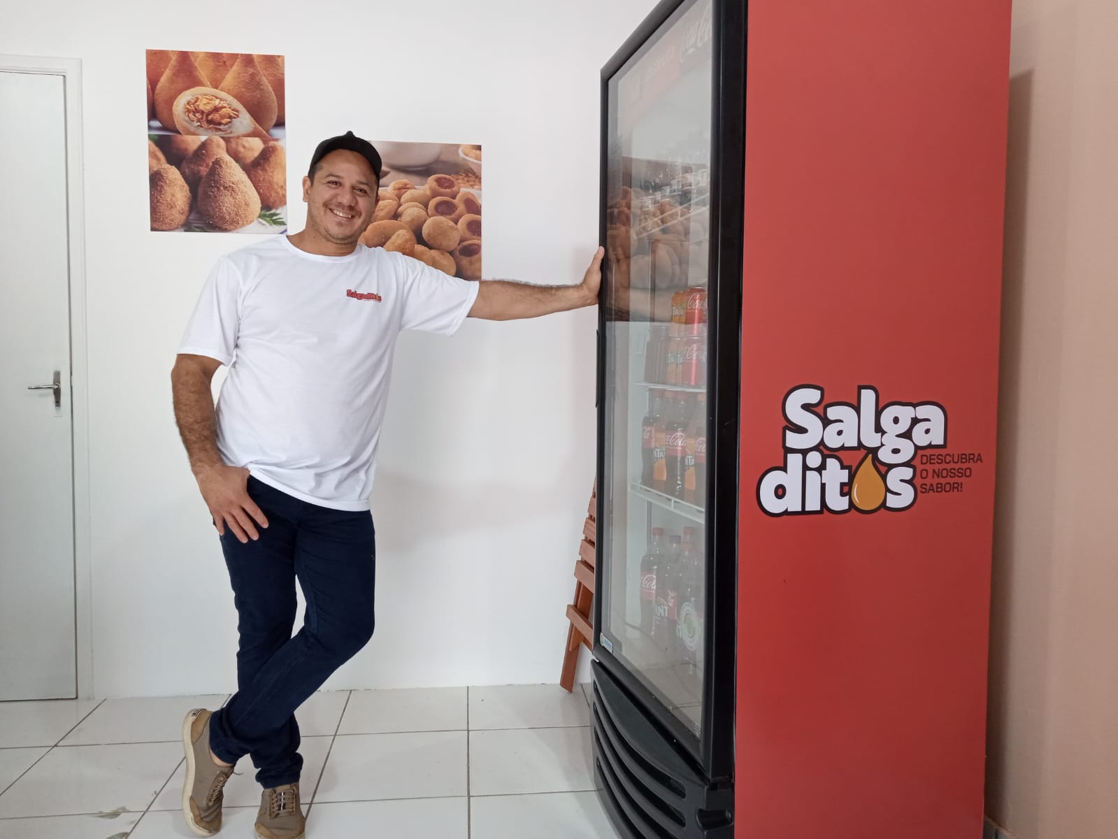 Salgaditos Um homem vestindo uma camiseta branca e calças escuras está de pé com uma mão apoiada em uma máquina de venda automática vermelha. A máquina tem o texto "Salgaditos: Descubra o Melhor Sabor" escrito nela. Há dois cartazes na parede atrás dele, ambos exibindo imagens de salgados. O chão é de azulejos e há uma porta fechada ao fundo.