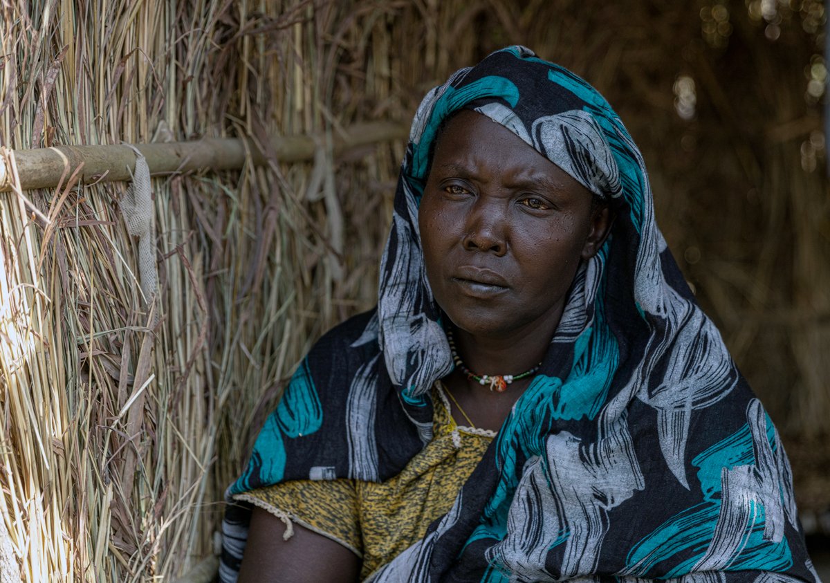Sudanese refugee Hawa A woman in front of a thatched shelter.