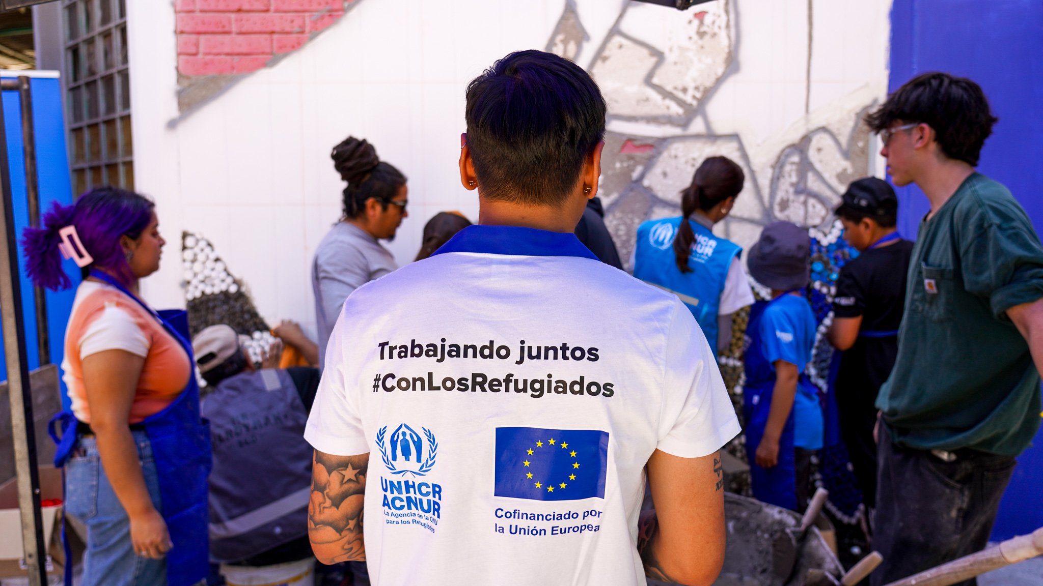 De espaldas un hombre que viste camiseta blanca con logotipos de ACNUR y la Unión Europera. Arriba se lee en color negro: Trabajando Juntos #ConLosRefugiados
