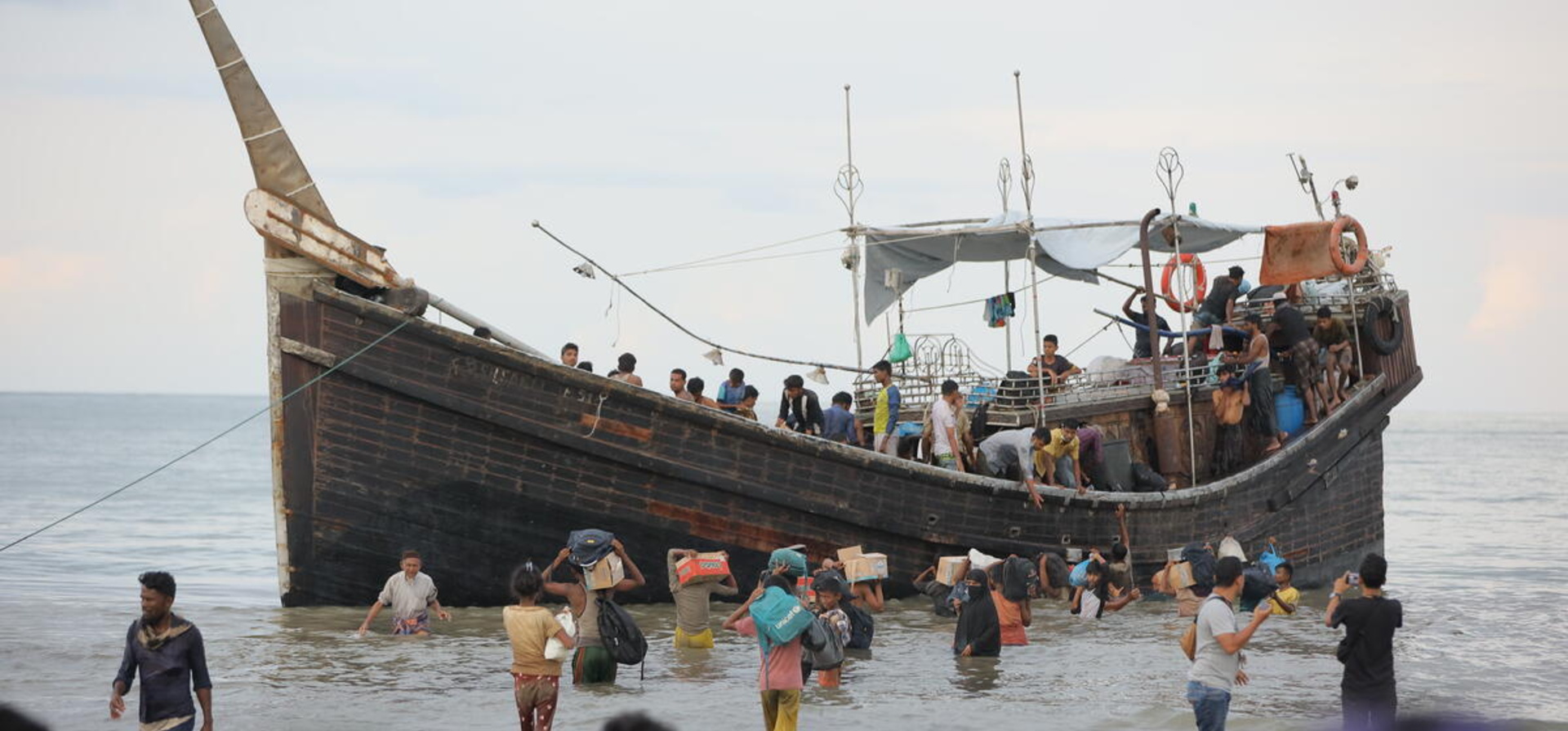 Un gran número de personas bajando de un barco; no han llegado a un muelle, así que muchas de ellas van cargando sus pertenencias con el agua hasta las rodillas.