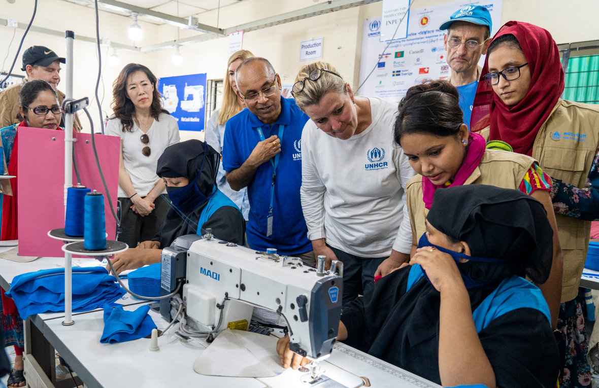 A group of people talk to a woman sitting at a sewing machine. Un grupo de personas habla con una mujer sentada frente a una máquina de coser.