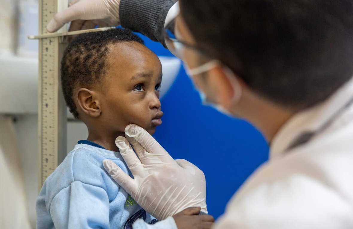 Un niño con sudadera azul claro frente a una regla mientras un hombre con mascarilla y guantes mide su estatura