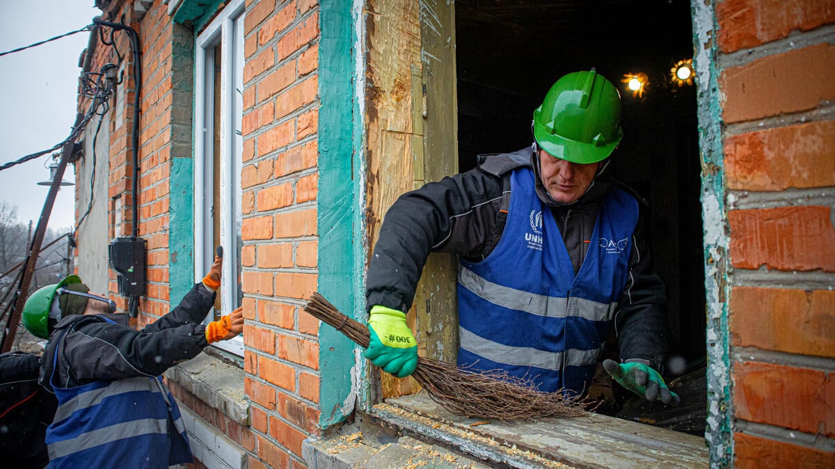 Dos hombres que llevan puestos cascos de construcción trabajan en la reparación de las ventanas de un vivienda hecha de ladrillo.