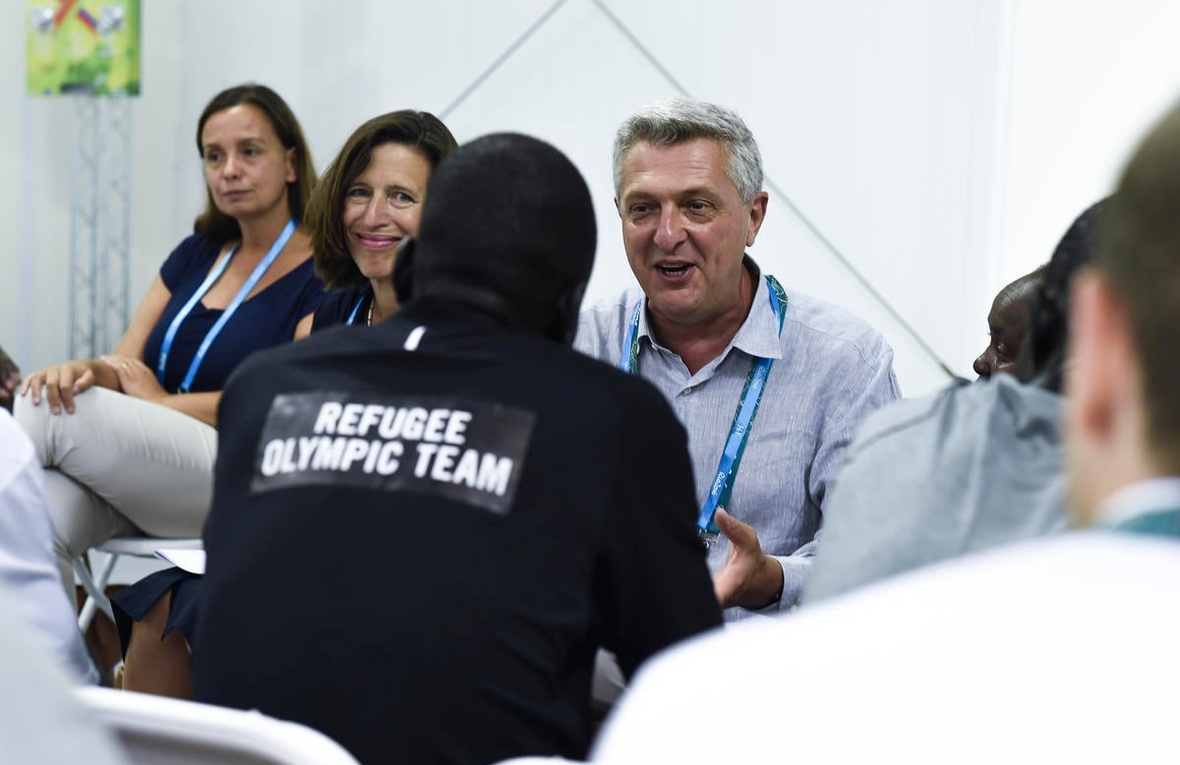Un hombre cano charla con un hombre que le da la espalda a la cámara y que lleva puesta una camiseta que dice "Equipo Olímpico de Atletas Refugiados". A lado del hombre cano hay dos mujeres; una de ellas le sonríe al integrante del Equipo Olímpico de Atletas Refugiados.