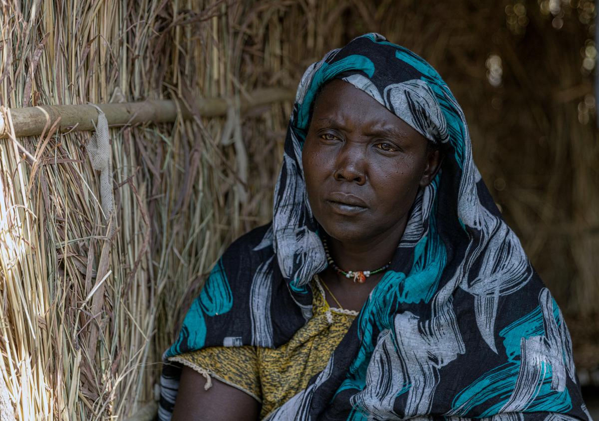 Sudanese refugee Hawa A woman in front of a thatched shelter.