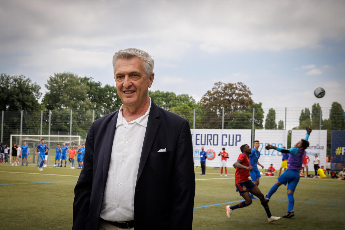 Un hombre cano en una cancha de fútbol sonríe a la cámara. Detrás suyo, dos equipos jugando.