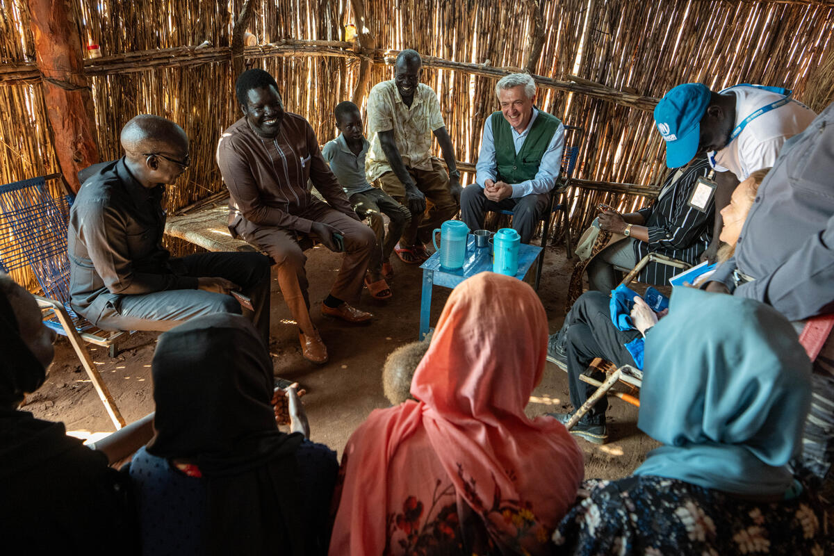 Un grupo de personas reunidas en un alojamiento hechizo; entre ellas, el Alto Comisionado de la ONU para los Refugiados, que es un hombre cano
