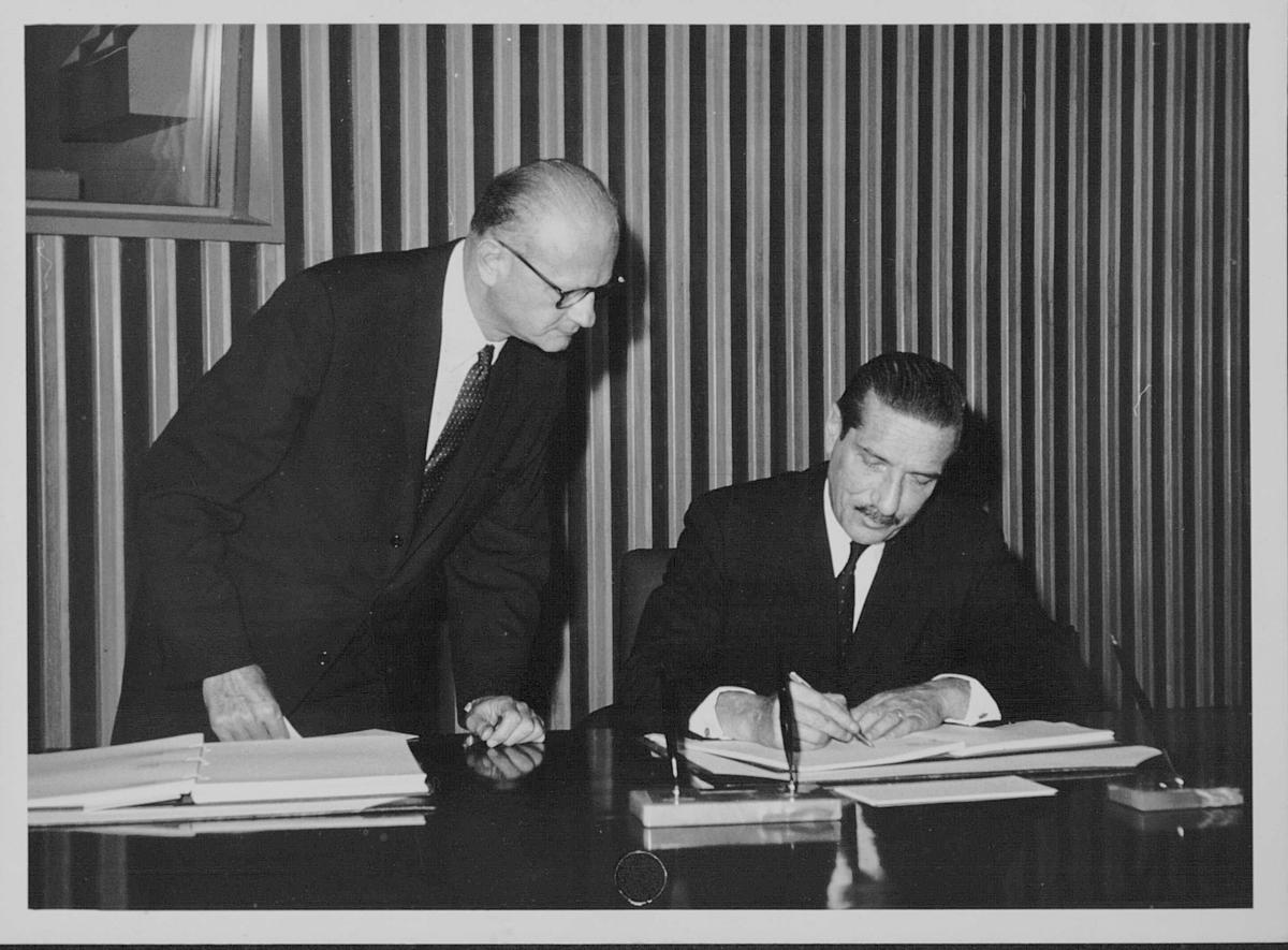 Imagen en blanco y negro. Dos hombres (uno, sentado; el otro, de pie a su lado). El hombre sentado, que aparece a la derecha, está firmando un documento; el otro observa.