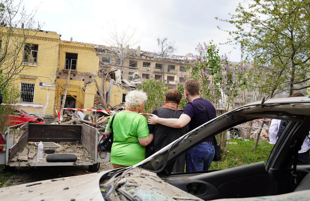 Tres personas fotografiadas de espaldas miran hacia unos edificios gravemente dañados y rodeados de escombros. Tres personas fotografiadas de espaldas miran hacia unos edificios gravemente dañados y rodeados de escombros.