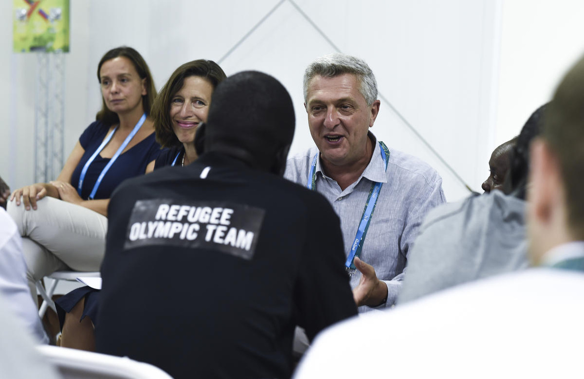 Un hombre cano charla con un hombre que le da la espalda a la cámara y que lleva puesta una camiseta que dice "Equipo Olímpico de Atletas Refugiados". A lado del hombre cano hay dos mujeres; una de ellas le sonríe al integrante del Equipo Olímpico de Atletas Refugiados.