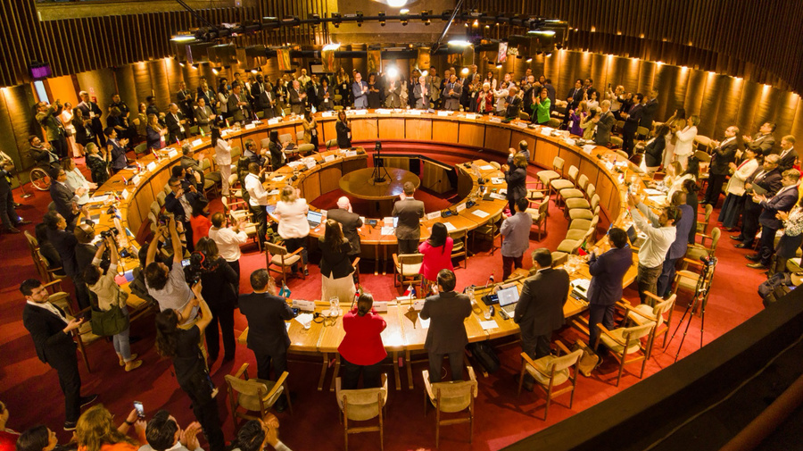 Panoraminc view of the conference room while delegates stand and clap their hands.