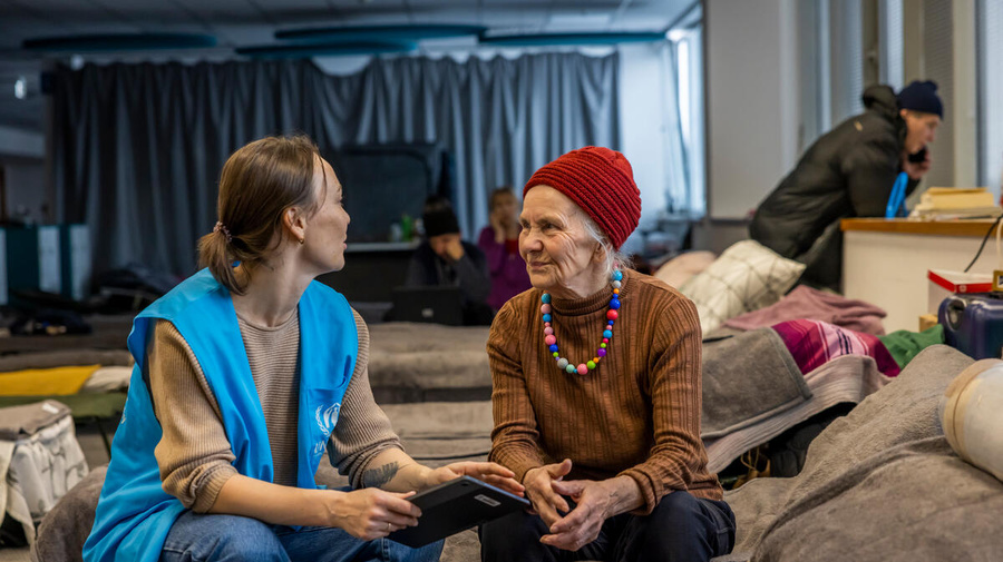 Sentadas sobre una cama en un albergue, una trabajadora de ACNUR charla con una mujer mayor.